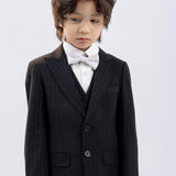 A young boy wearing glasses and KIKI and JOJO’s Kids Suit Pants – Black (3-14 Years), Classic Fit, Wrinkle-Free, with a white shirt and bow tie, stands against a plain light background, looking down.