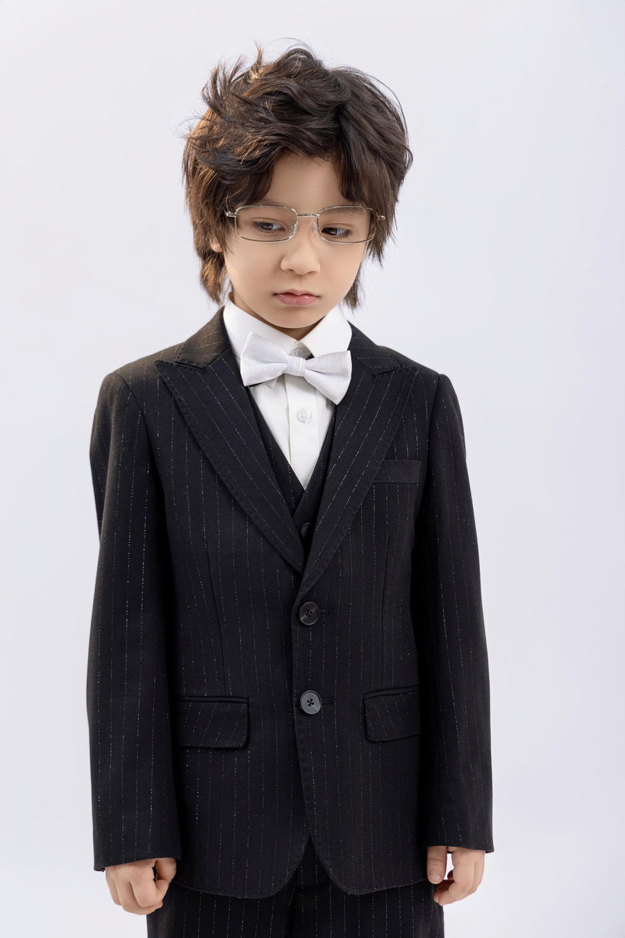 A young boy wearing glasses and KIKI and JOJO’s Kids Suit Pants – Black (3-14 Years), Classic Fit, Wrinkle-Free, with a white shirt and bow tie, stands against a plain light background, looking down.
