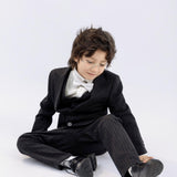A young boy wearing KIKI and JOJO's Kids Suit Pants – Black (Classic Fit, Wrinkle-Free, ages 3-14) with a white bow tie sits on the floor and touches his shoe against a plain white background.