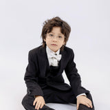 A young boy with glasses, dressed in a black suit and bow tie with KIKI and JOJO Kids Suit Pants – Black (3-14 Years), Classic Fit, Wrinkle-Free, sits on the floor against a plain light background.