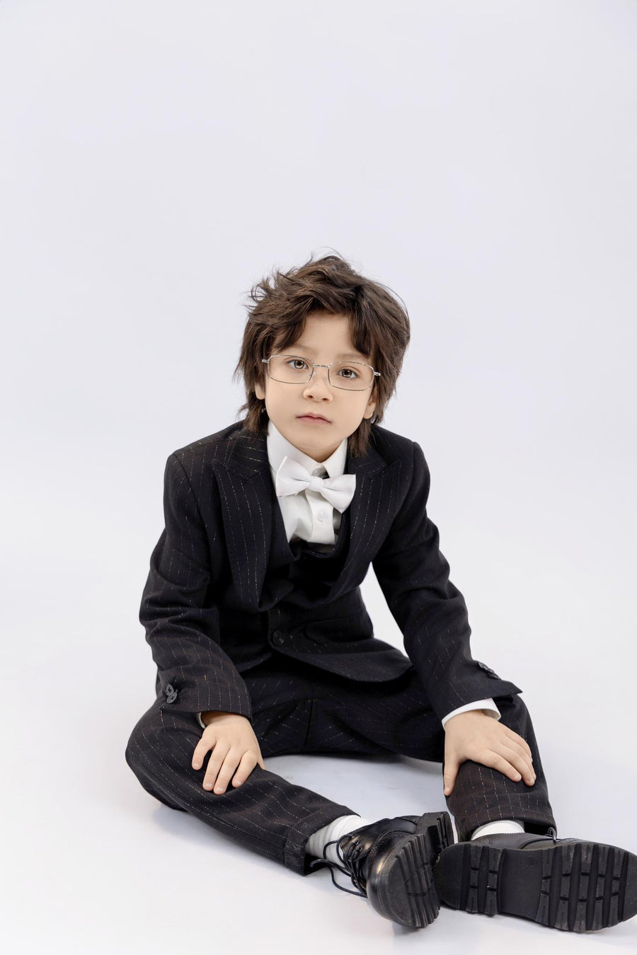A young boy with glasses, dressed in a black suit and bow tie with KIKI and JOJO Kids Suit Pants – Black (3-14 Years), Classic Fit, Wrinkle-Free, sits on the floor against a plain light background.