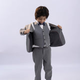 A young boy in a KIKI and JOJO Kids Suit Jacket – Gray (3-14 Years), classic fit and wrinkle-free, stands looking down as he holds open his jacket, paired with black shoes.