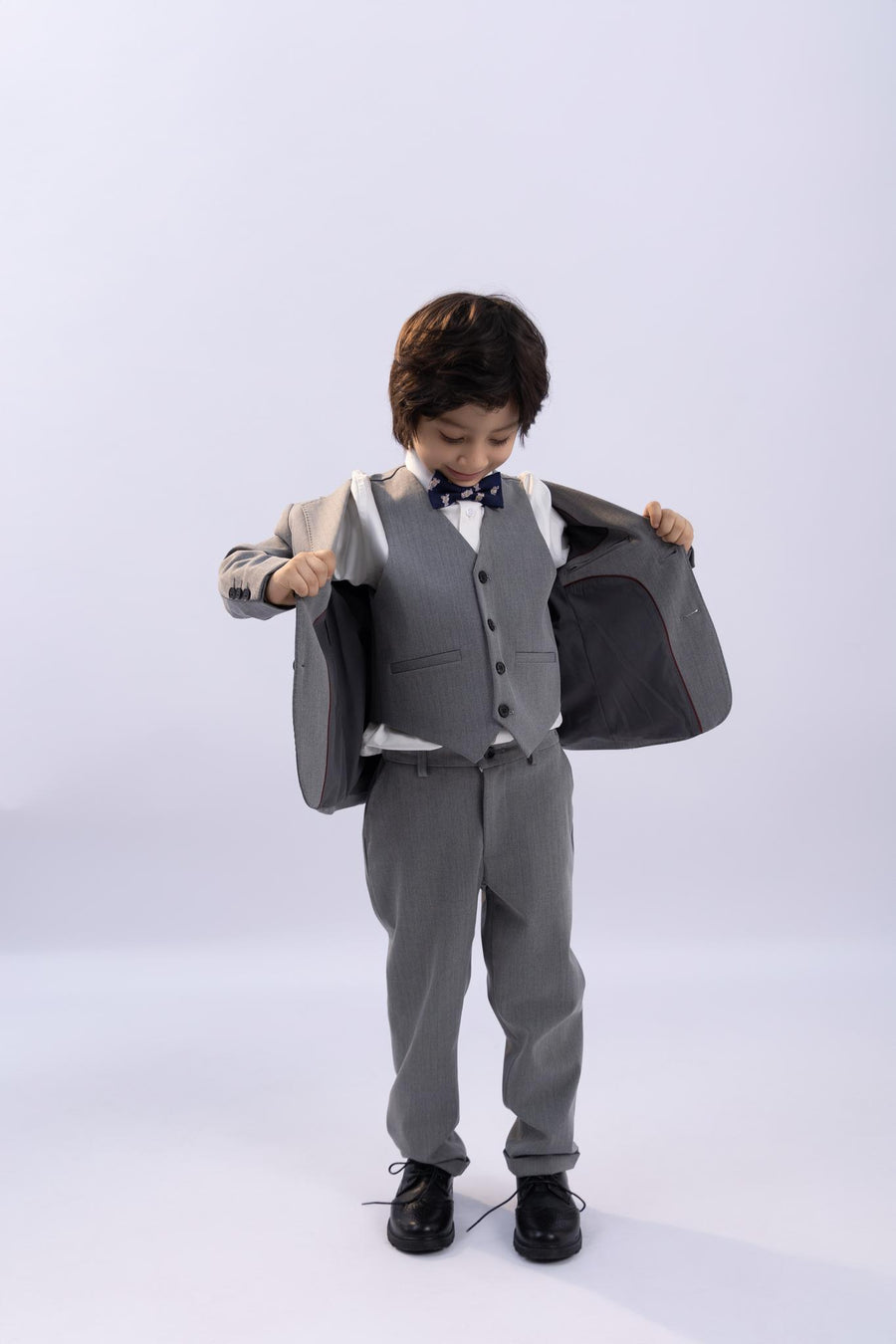 A young boy in a KIKI and JOJO Kids Suit Jacket – Gray (3-14 Years), classic fit and wrinkle-free, stands looking down as he holds open his jacket, paired with black shoes.