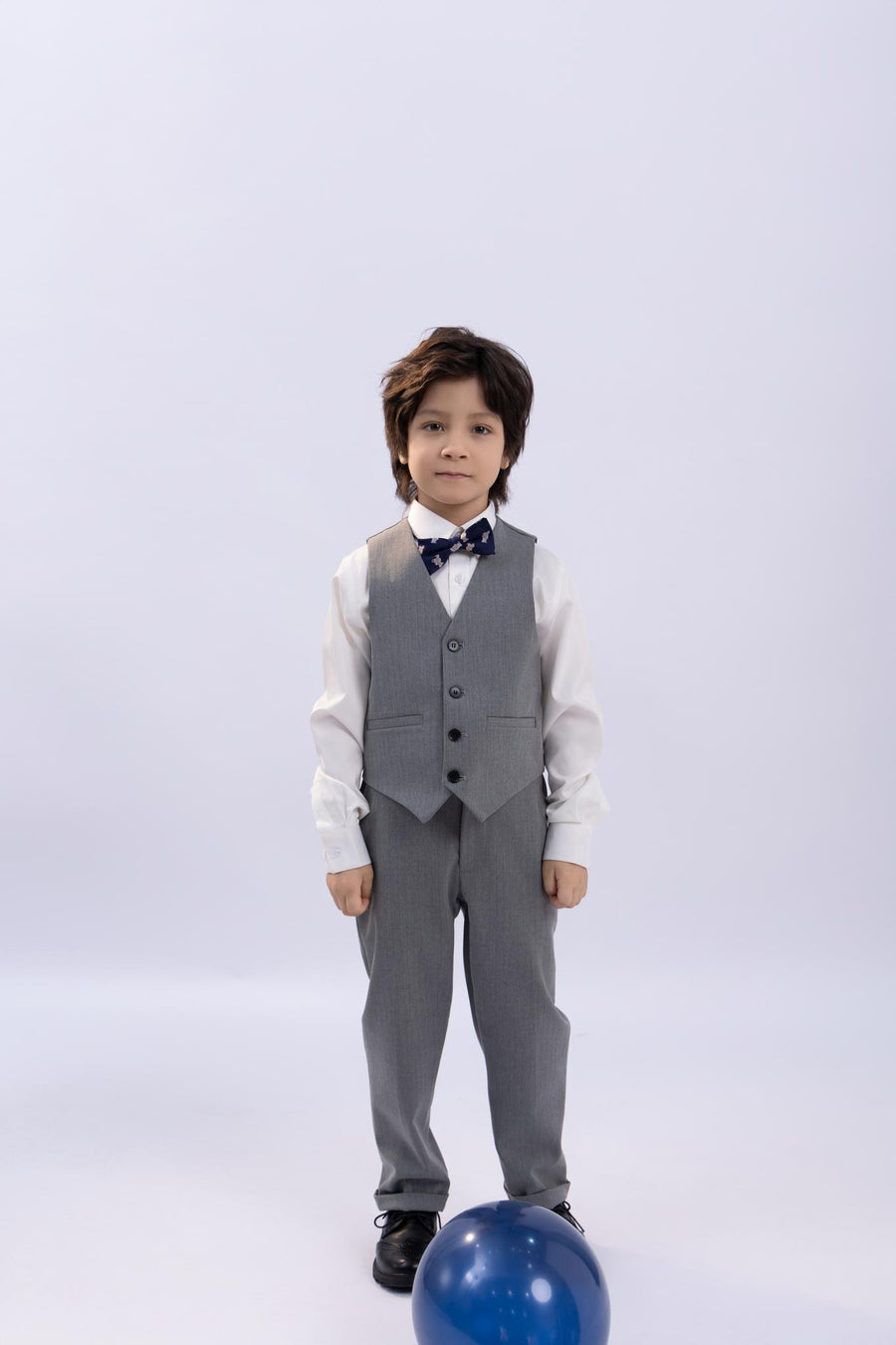 A young boy wearing the KIKI and JOJO Kids Suit – Gray 3-Piece Set (Classic Fit, Wrinkle-Free, ages 3-14) stands on a white background with a blue balloon on the floor in front of him.