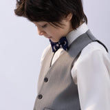 A young boy in a white shirt, grey vest, navy bow tie, and KIKI and JOJO Kids Suit Pants – Gray (Classic Fit, Wrinkle-Free) looks downward against a plain light background.