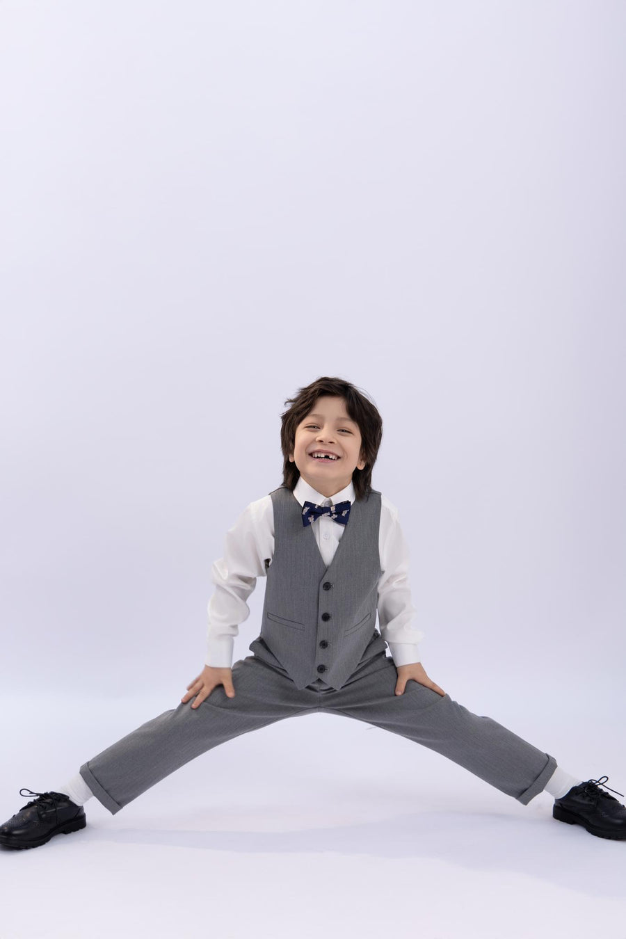 A young boy beams in a KIKI and JOJO Kids Suit – Gray 3-Piece Set (Classic Fit, Wrinkle-Free) as he does a wide-leg split against a plain light background.