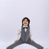A young boy in a gray suit and bow tie does a split, smiling against a white background—highlighting the classic fit and wrinkle-free fabric of KIKI and JOJO Kids Suit Pants – Gray (3-14 Years).