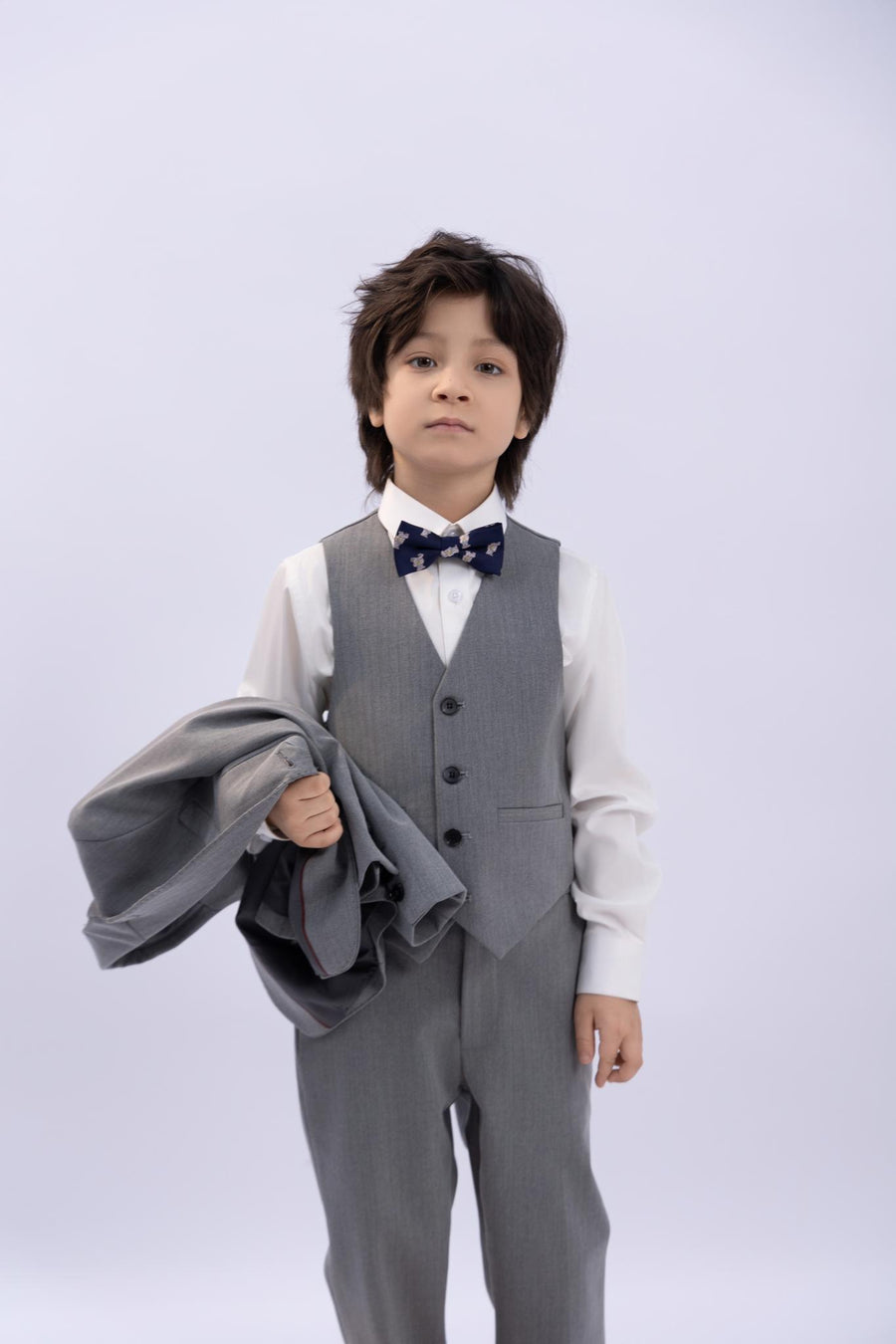 A young boy wears the KIKI and JOJO Kids Suit – Gray 3-Piece Set (3-14 Years), a classic fit wrinkle-free ensemble with a white shirt and bow tie, holding his suit jacket against a plain light background.