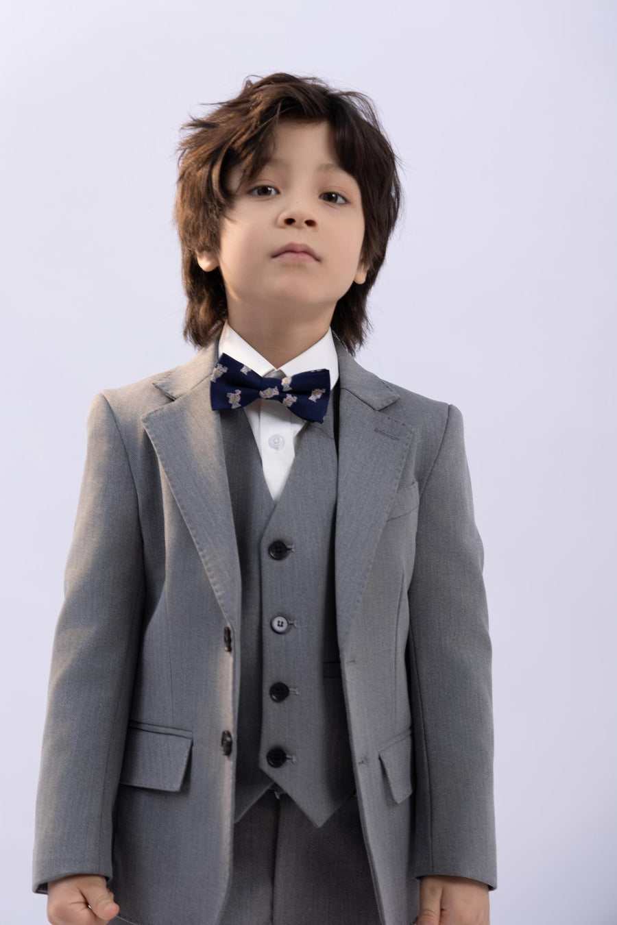 A young boy models the KIKI and JOJO Kids Suit – Gray 3-Piece Set (3-14 Years), featuring a classic fit, wrinkle-free fabric, white shirt, and dark patterned bow tie against a plain light background.
