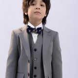 A young boy models the KIKI and JOJO Kids Suit Jacket – Gray (3-14 Years), Classic Fit, Wrinkle-Free, paired with a vest, white shirt, and patterned bow tie against a plain light background.