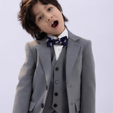 A young child wearing the KIKI and JOJO Kids Suit Jacket – Gray (3-14 Years) – Classic Fit – Wrinkle-Free, paired with a white shirt and patterned bow tie, stands with head tilted and mouth open against a plain light background.