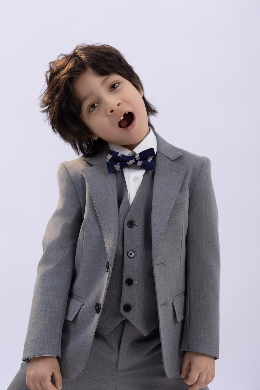 A young child wearing the KIKI and JOJO Kids Suit Jacket – Gray (3-14 Years) – Classic Fit – Wrinkle-Free, paired with a white shirt and patterned bow tie, stands with head tilted and mouth open against a plain light background.