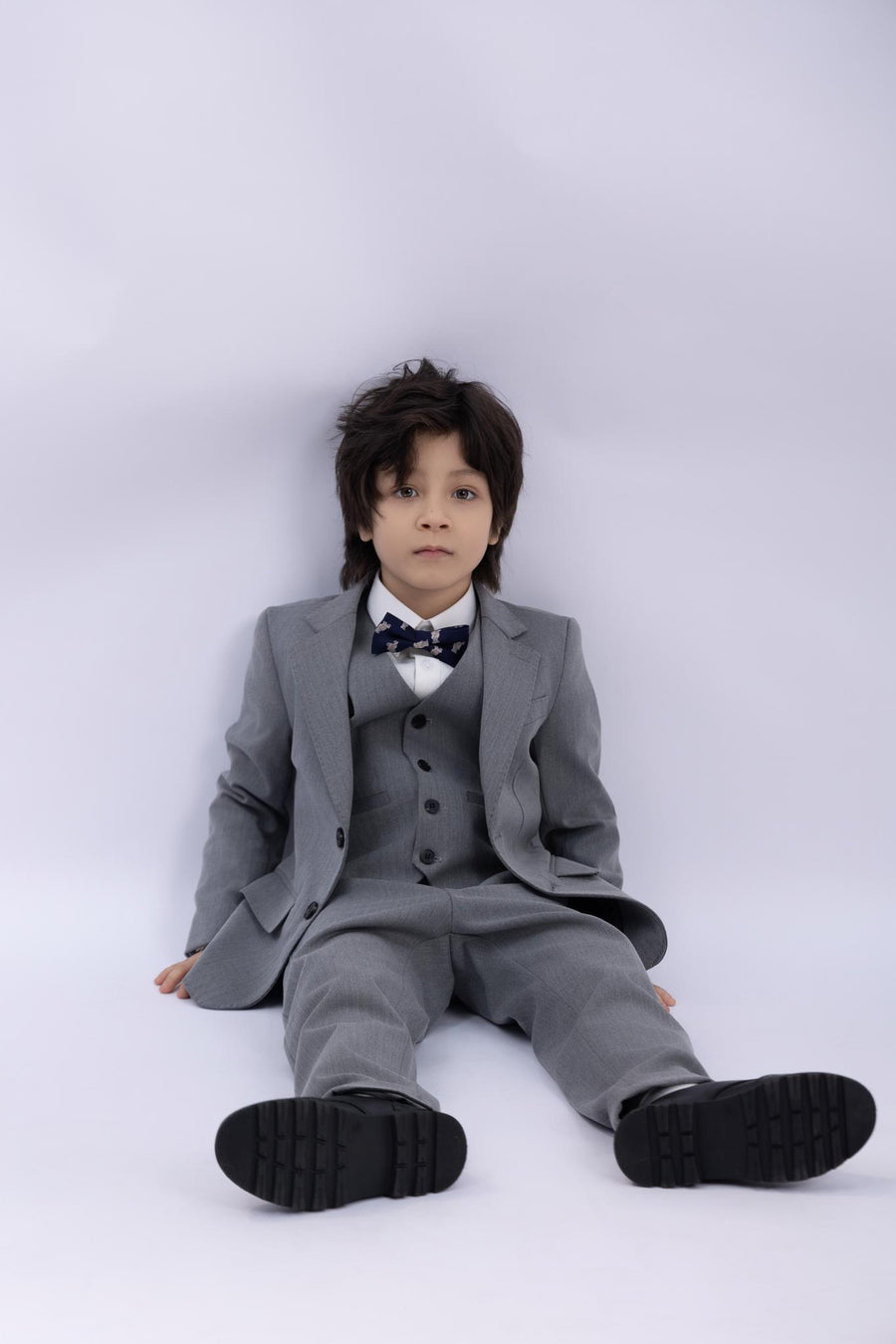A young boy in a KIKI and JOJO Kids Suit – Gray 3-Piece Set (3-14 Years), classic fit and wrinkle-free, sits on the floor against a plain light background, looking at the camera.