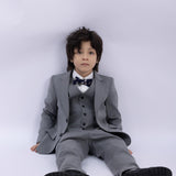 A young boy sits on the floor against a white background wearing a KIKI and JOJO Kids Suit Jacket – Gray (3-14 Years), Classic Fit, Wrinkle-Free, paired with a bow tie.
