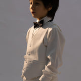 A young boy wearing the KIKI and JOJO Kids Suit Vest – White (3-14 Years), classic fit and wrinkle-free, paired with a white shirt and black bow tie, stands with his hands in his pockets, gazing to the side.