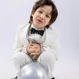 A young child in a KIKI and JOJO Kids Suit Vest – White (3-14 Years), Classic Fit, Wrinkle-Free, with a black bow tie, sits crouched on the floor beside a silver balloon against a plain light background.