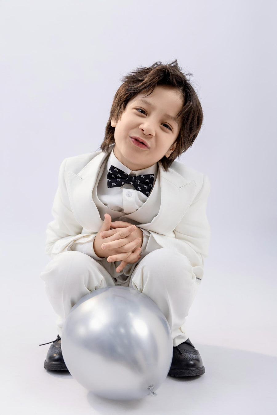 A young child in a KIKI and JOJO Kids Suit Vest – White (3-14 Years), Classic Fit, Wrinkle-Free, with a black bow tie, sits crouched on the floor beside a silver balloon against a plain light background.