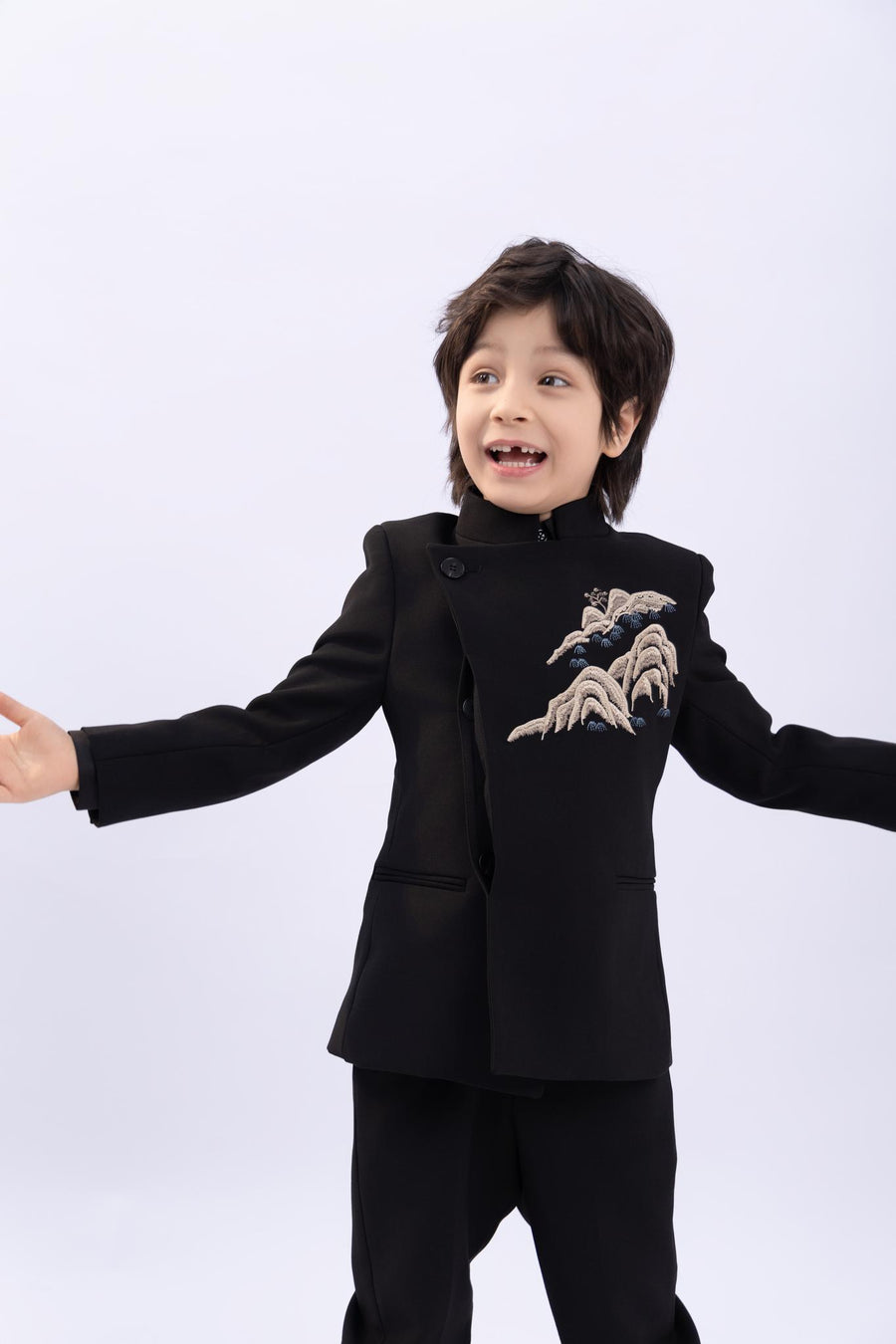 A young boy smiles with arms wide open, wearing the KIKI and JOJO Kids Suit Jacket – Black (3-14 Years), a slim-fit, wrinkle-free jacket, standing against a plain background.