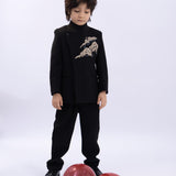 A young boy wears the KIKI and JOJO Kids Suit Jacket – Black (3-14 Years), a slim-fit, wrinkle-free design with a Classic Mandarin collar, as he stands on a white background looking down at two red balloons.