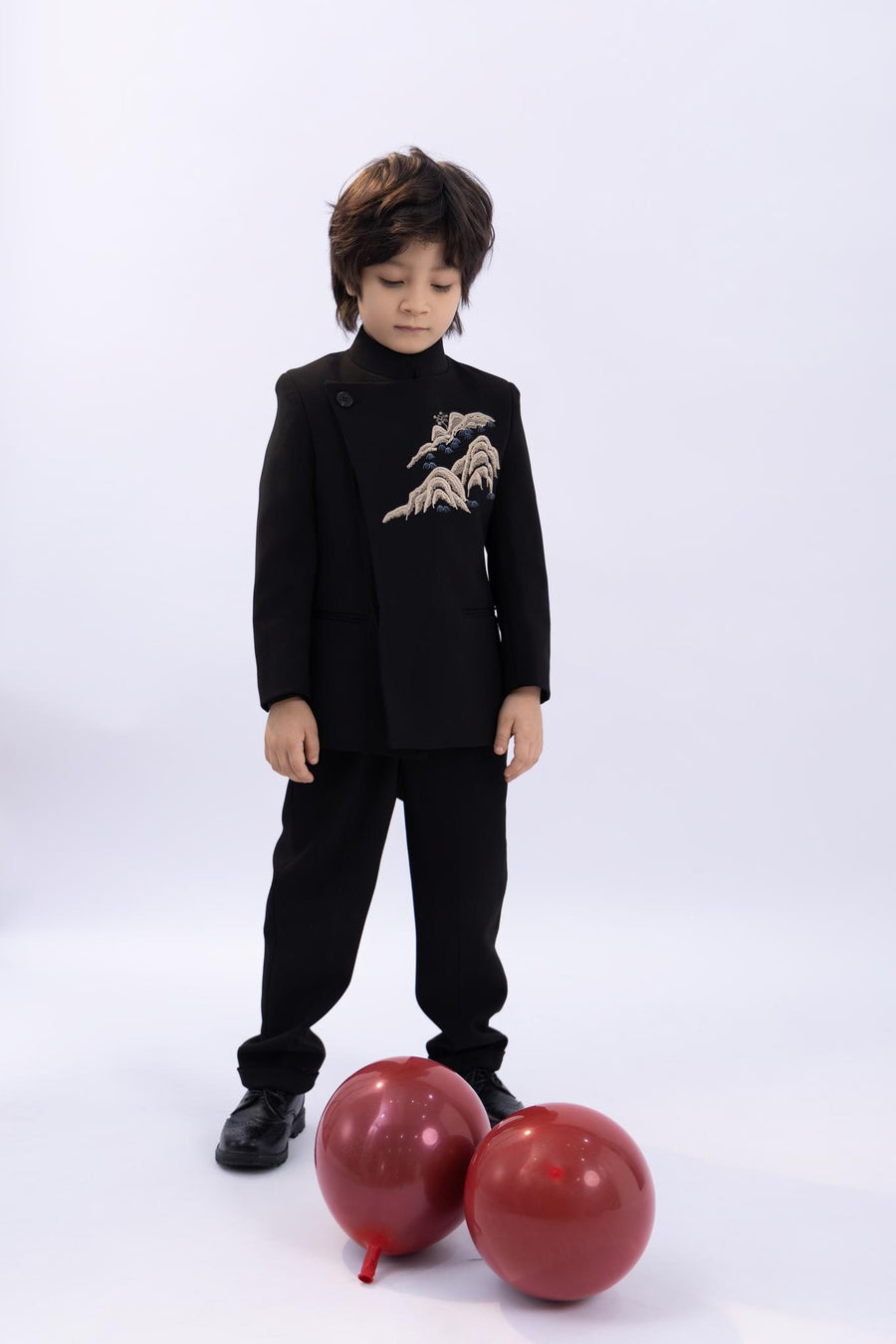 A young boy wears the KIKI and JOJO Kids Suit Jacket – Black (3-14 Years), a slim-fit, wrinkle-free design with a Classic Mandarin collar, as he stands on a white background looking down at two red balloons.