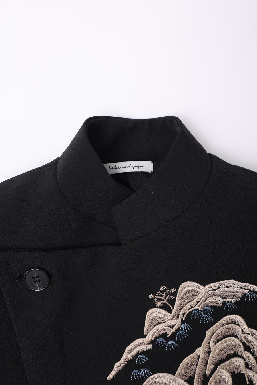 Close-up of the KIKI and JOJO Kids Suit Jacket – Black (3-14 Years), a slim-fit, wrinkle-free design featuring a Classic Mandarin collar, black button, mountain embroidery, and the brand’s inner label.