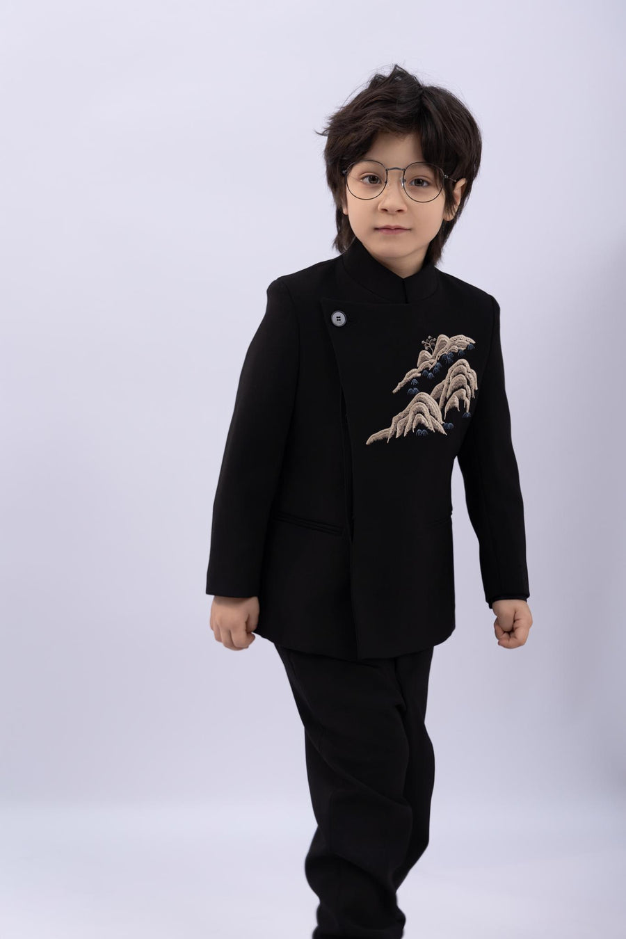 A young child with dark hair and glasses models the KIKI and JOJO Kids Suit – Black 2-Piece Set (3-14 Years), a slim fit, wrinkle-free outfit with a Mandarin collar and embroidered chest, set against a plain light background.