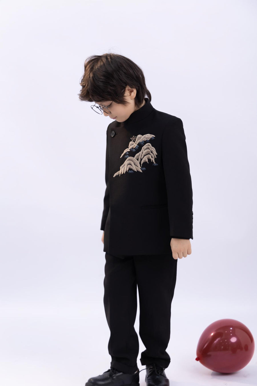 A young boy wears the KIKI and JOJO Kids Suit Jacket in black (slim-fit, wrinkle free), featuring embroidered detail and a classic Mandarin collar, as he stands looking down next to a red balloon on a white background.