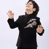 A young boy wears the KIKI and JOJO Kids Suit Jacket – Black (3-14 Years), a slim-fit, wrinkle-free jacket with embroidered patterns and a Classic Mandarin collar. Smiling, he stands with hands raised as a red balloon lies on the floor beside him.