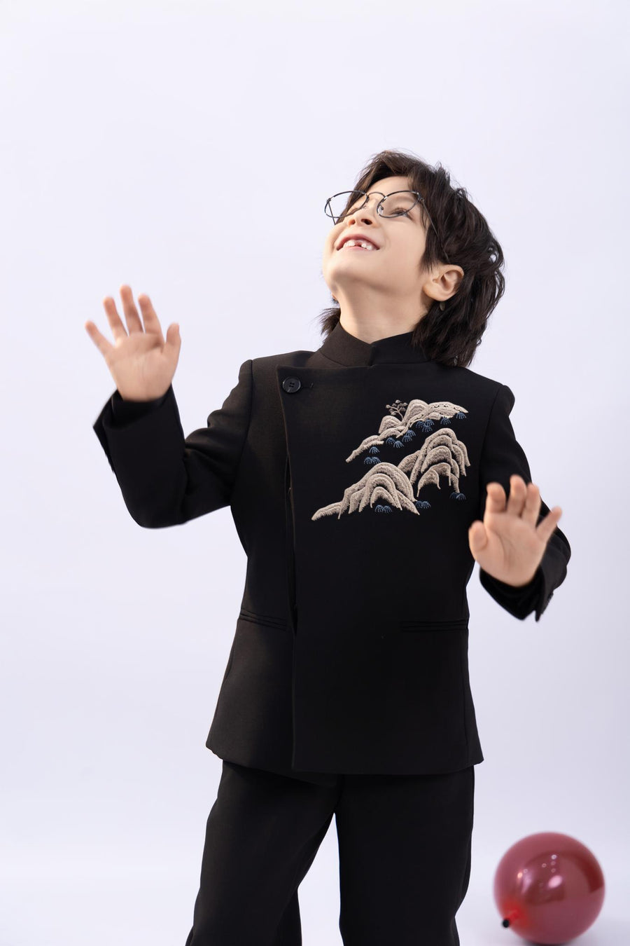 A young boy wears the KIKI and JOJO Kids Suit Jacket – Black (3-14 Years), a slim-fit, wrinkle-free jacket with embroidered patterns and a Classic Mandarin collar. Smiling, he stands with hands raised as a red balloon lies on the floor beside him.