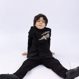 A young boy wears the KIKI and JOJO Kids Suit Jacket – Black (3-14 Years), a slim-fit, wrinkle-free suit with a classic Mandarin collar, as he sits on the floor against a plain white background.
