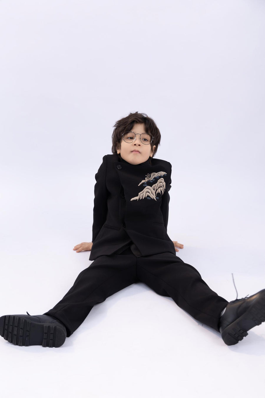 A young boy wears the KIKI and JOJO Kids Suit Jacket – Black (3-14 Years), a slim-fit, wrinkle-free suit with a classic Mandarin collar, as he sits on the floor against a plain white background.