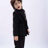 A young boy with glasses wears the KIKI and JOJO Kids Suit Jacket – Black (3-14 Years), a slim-fit, wrinkle-free jacket with a classic mandarin collar, as he stands against a plain light background looking surprised.