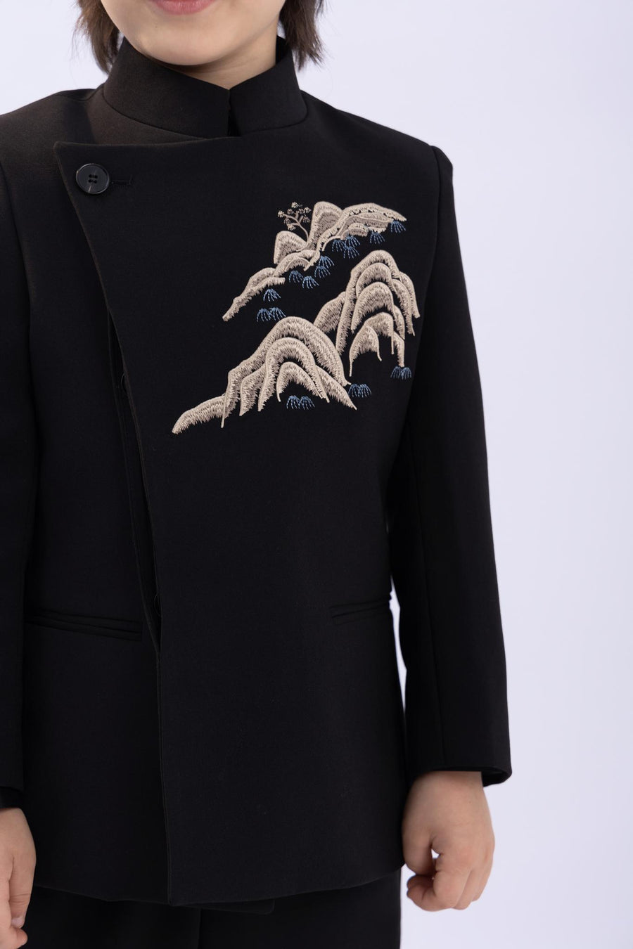 A child models the KIKI and JOJO Kids Suit – Black 2-Piece Set (3-14 Years), a slim fit, wrinkle-free jacket with a Mandarin collar and embroidered mountain detail, set against a plain light background.