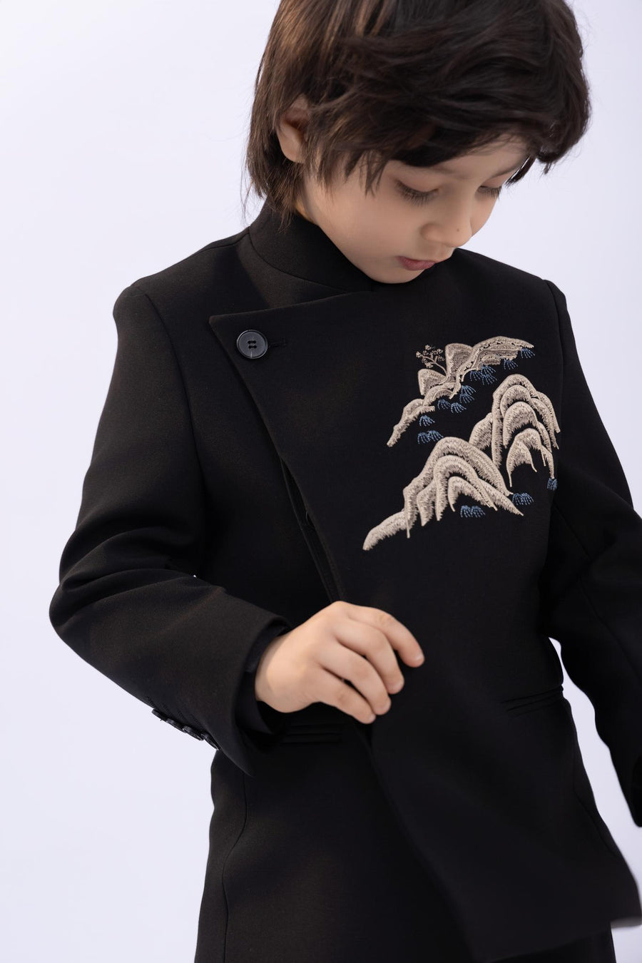 A child wearing the KIKI and JOJO Kids Suit – Black 2-Piece Set (slim fit, wrinkle-free) for ages 3-14 adjusts the front of their jacket.