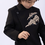 A child wearing the KIKI and JOJO Kids Suit Jacket – Black (3-14 Years), a slim-fit, wrinkle-free blazer with a classic Mandarin collar and embroidered mountain design, looks down while adjusting the front.