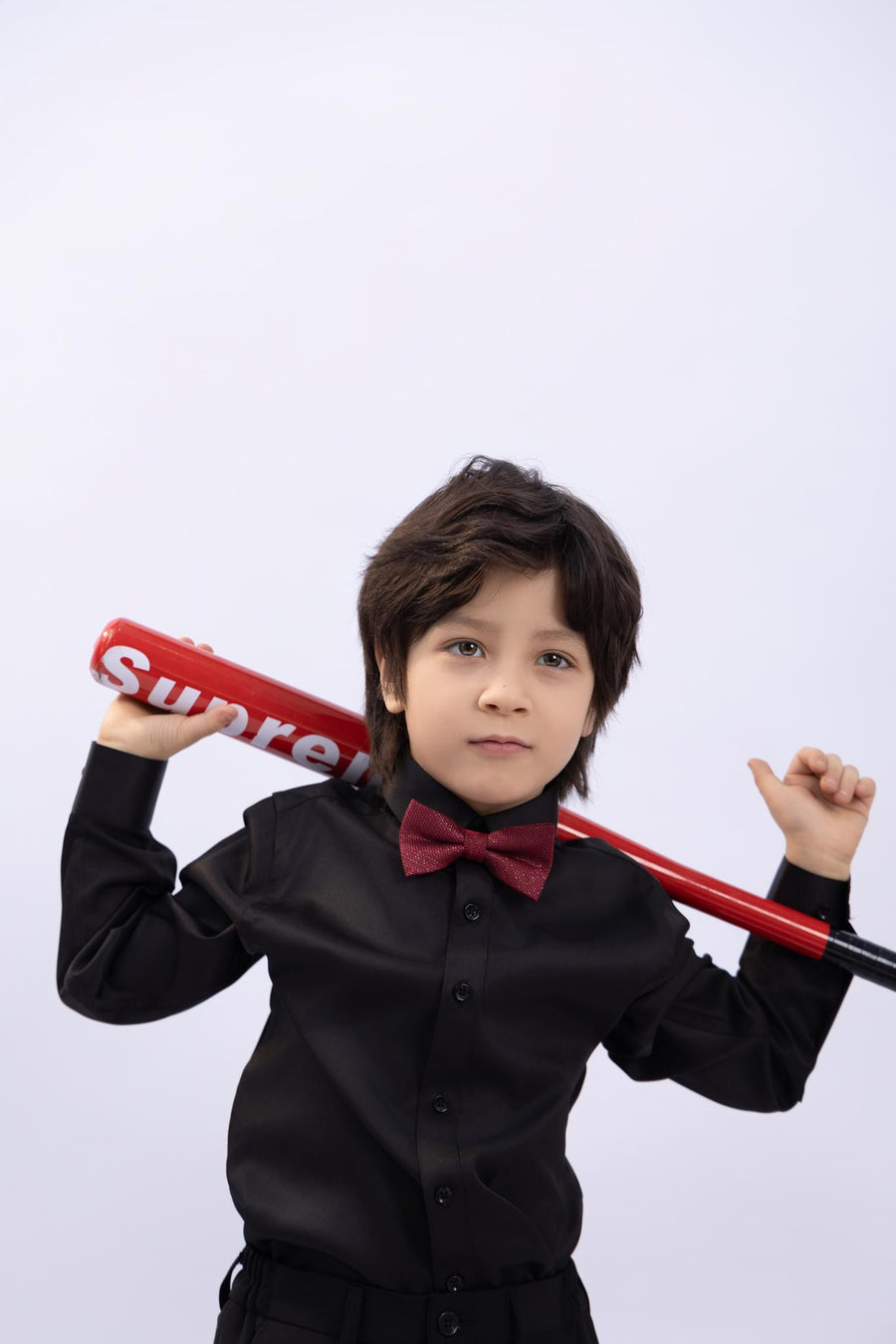 A young boy models the KIKI and JOJO Kids Dress Shirt – Black Long Sleeve (3-14 Years), classic fit, with a red bow tie and bat, making a stylish statement against a plain white background.