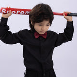 A young boy wears the KIKI and JOJO Kids Dress Shirt – Black Long Sleeve (Classic Fit, 3-14 Years) with a red bow tie, holding a red Supreme baseball bat across his shoulders against a light background.