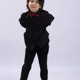 A young boy stands with his hands behind his back, wearing the KIKI and JOJO Kids Dress Shirt – Black Long Sleeve (Classic Fit, 3-14 Years) with black pants, black shoes, and a red bow tie against a plain light background.