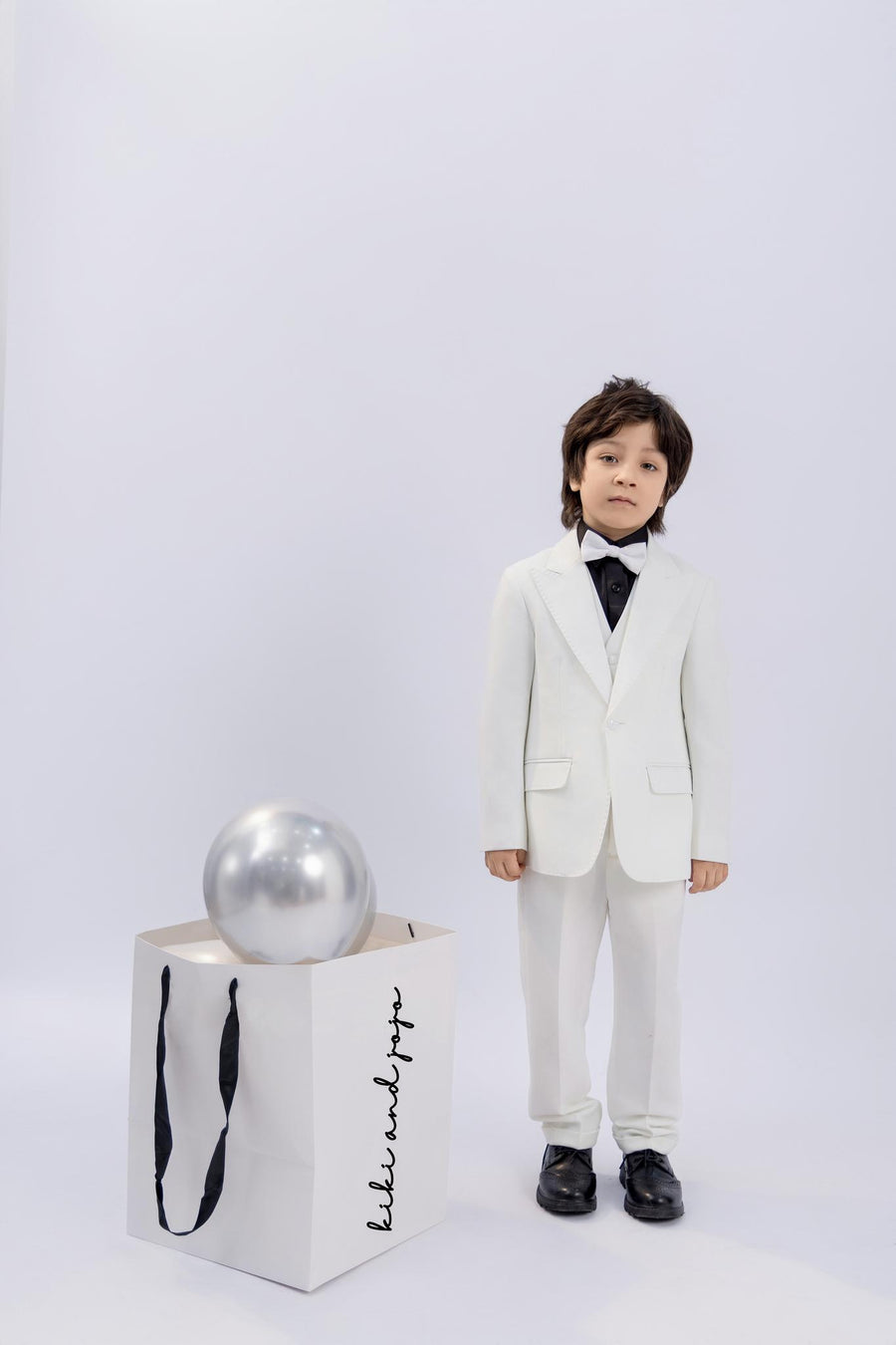 A young boy wears the KIKI and JOJO Kids Dress Shirt – Black Long Sleeve (Classic Fit, 3-14 Years) with a white suit and black shoes, standing beside a large gift bag holding a silver balloon against a plain light background.