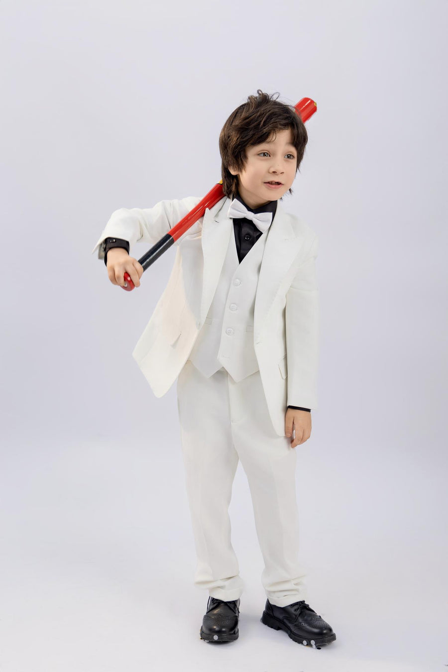 A young boy wears the KIKI and JOJO Kids Dress Shirt – Black Long Sleeve (3-14 Years) – Classic Fit, paired with a white suit and holding a red baseball bat over his shoulder against a plain light background—a timeless look.