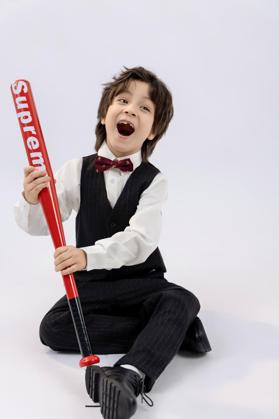 A boy wears a KIKI and JOJO Kids Dress Shirt – White Long Sleeve (Classic Fit), sitting on the floor, smiling with mouth open, holding a red "Supreme" baseball bat against a plain white background.