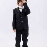 A young boy wearing the KIKI and JOJO Kids Dress Shirt – White Long Sleeve (Classic Fit, ages 3-14) stands against a plain background in a black suit and bow tie, covering one eye with his hand.