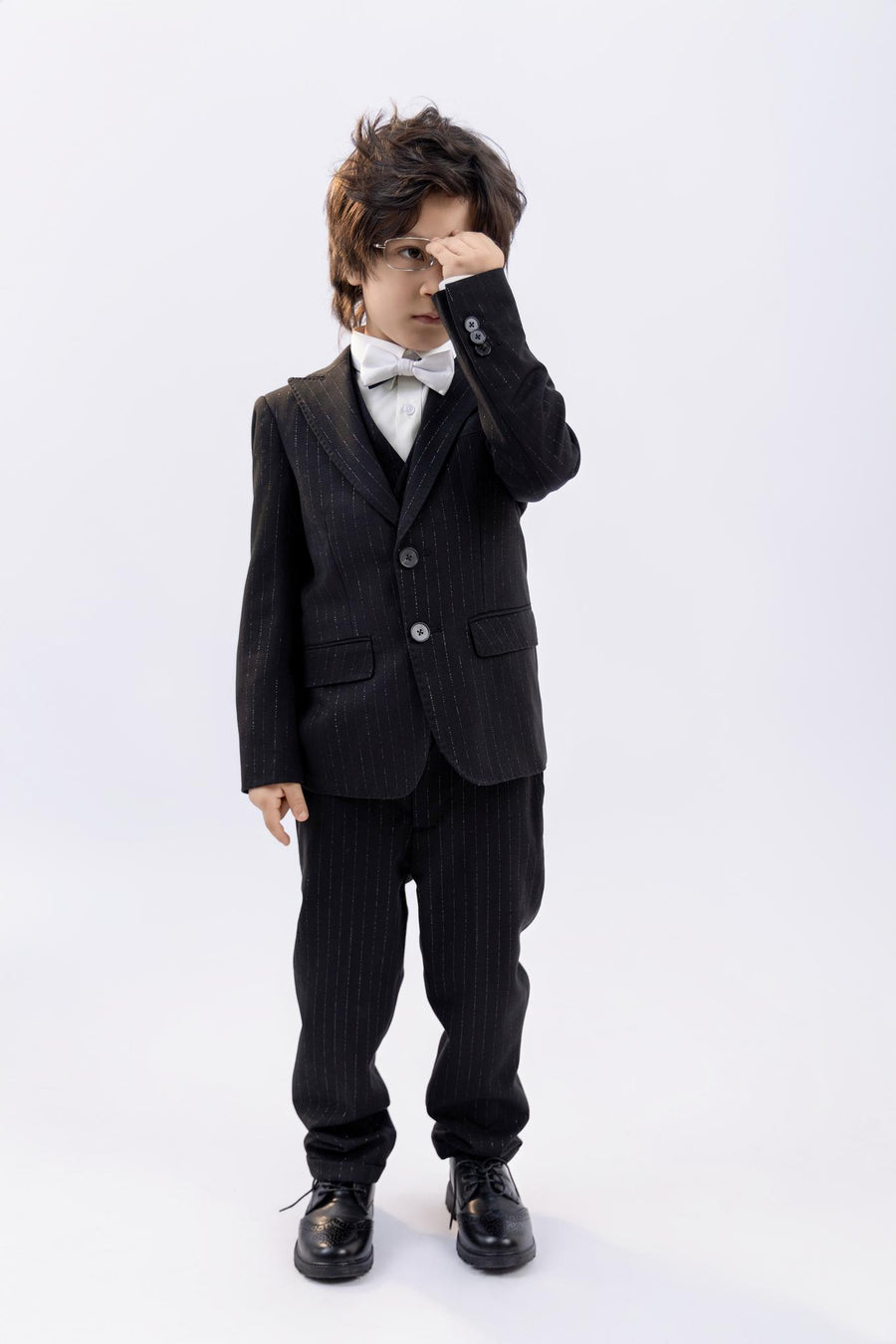A young boy wearing the KIKI and JOJO Kids Dress Shirt – White Long Sleeve (Classic Fit, ages 3-14) stands against a plain background in a black suit and bow tie, covering one eye with his hand.