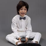 A young boy wears the KIKI and JOJO Kids Dress Shirt – White Long Sleeve (Classic Fit, 3-14 Years), paired with white pants and a bow tie, sitting cross-legged on the floor, holding a smartphone against a plain gray background.