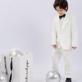 A young boy wears the KIKI and JOJO Kids Dress Shirt – White Long Sleeve (3-14 Years), classic fit, standing by silver balloons and a white gift bag with black handles against a plain background.