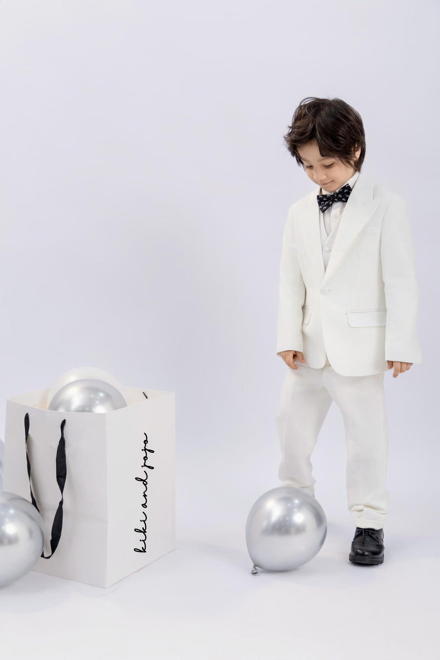 A young boy wears the KIKI and JOJO Kids Dress Shirt – White Long Sleeve (3-14 Years), classic fit, standing by silver balloons and a white gift bag with black handles against a plain background.