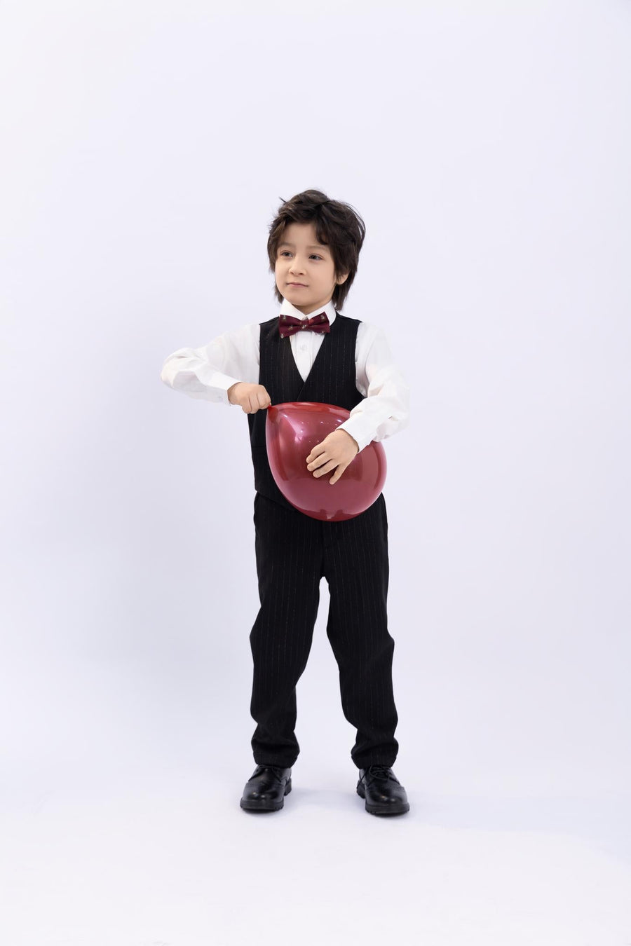 A young boy in a formal suit wears the KIKI and JOJO Kids Bow Tie – Burgundy Bear, a pre-tied adjustable satin bow tie, holding a red heart-shaped balloon against a plain white background.