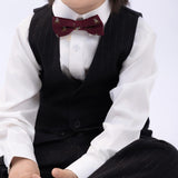 A young child in a black pinstriped suit and white shirt wears the KIKI and JOJO Kids Bow Tie – Burgundy Bear – Pre-Tied Satin, sitting cross-legged with mouth open against a plain background.