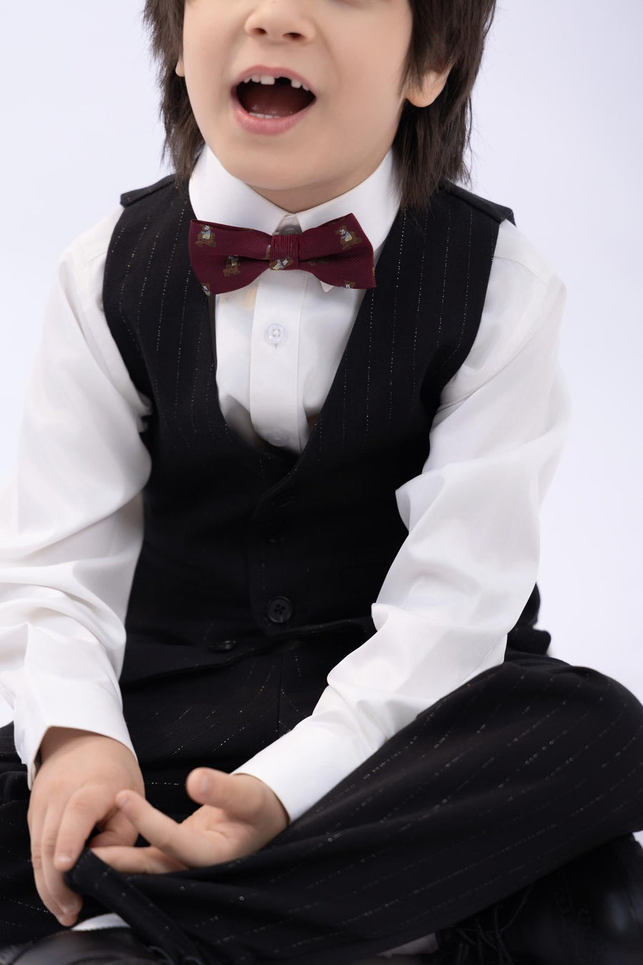 A young child in a black pinstriped suit and white shirt wears the KIKI and JOJO Kids Bow Tie – Burgundy Bear – Pre-Tied Satin, sitting cross-legged with mouth open against a plain background.
