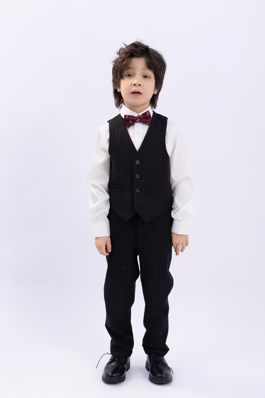 A young boy stands facing forward against a plain white background, wearing a white shirt, dark vest and pants, dress shoes, and the KIKI and JOJO Kids Bow Tie – Burgundy Bear – Pre-Tied Satin – Adjustable.
